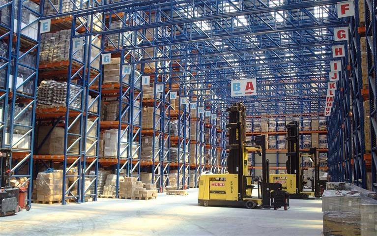 Interior of a large warehouse with tall shelves filled with boxes and yellow forklifts in the aisle.