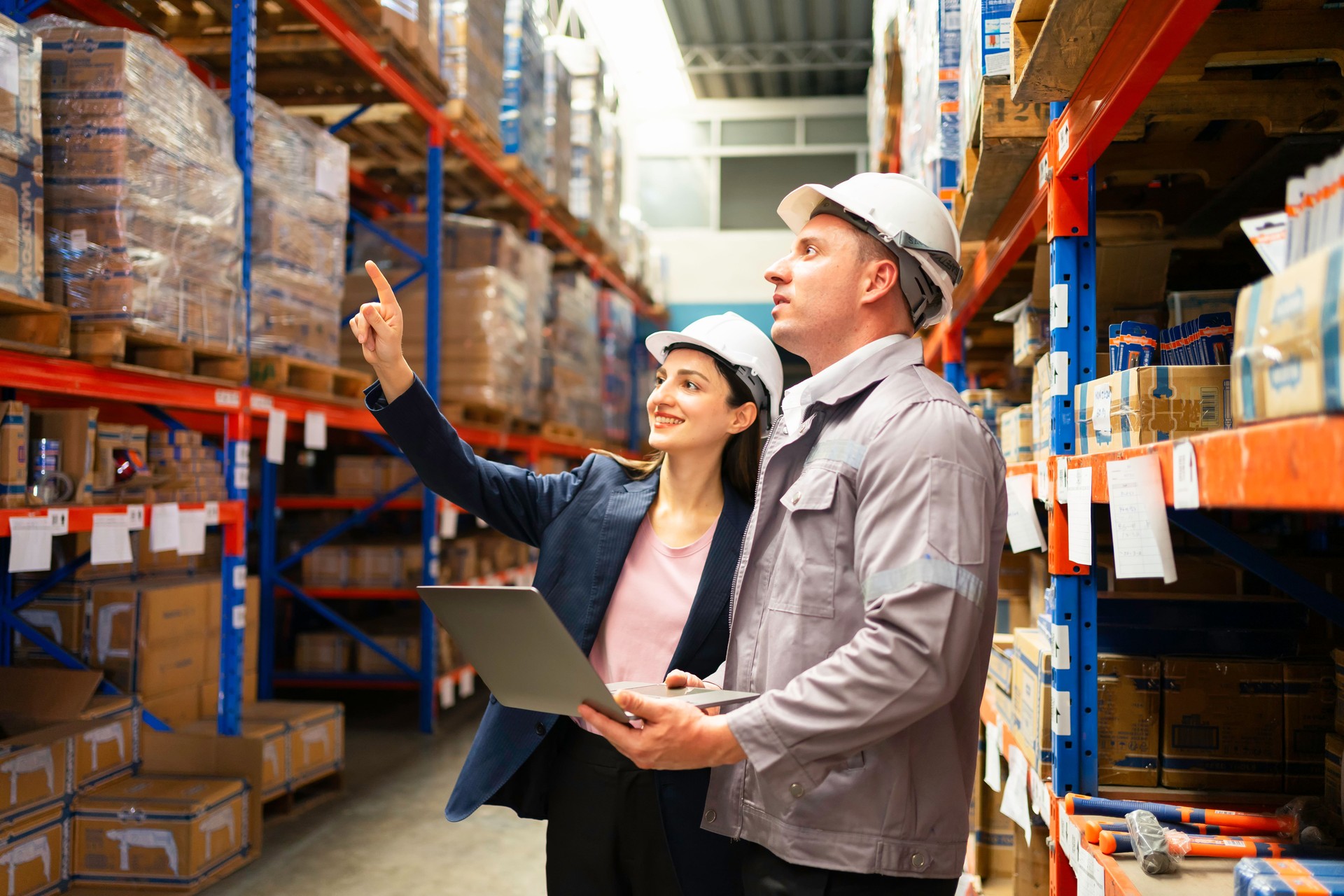 Two professionals inspect inventory in a warehouse, discussing operations and logistics while using a laptop for data analysis.